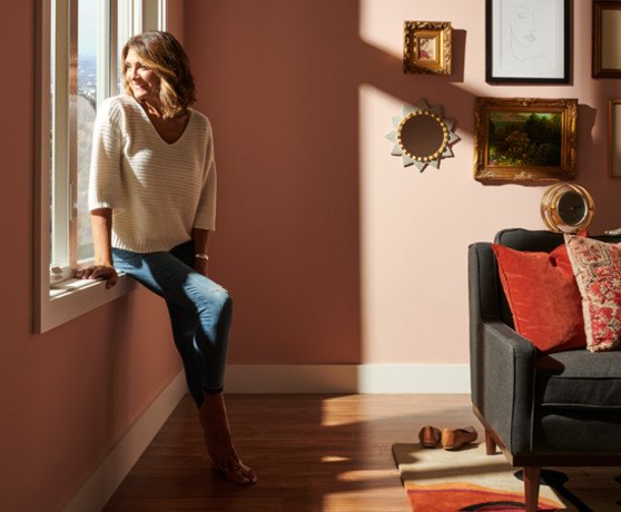 A woman sitting on the sill of a V300 window to the left. A peach colored room with various pictures on the wall
