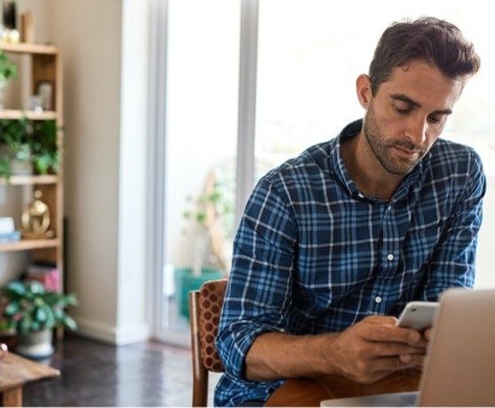 Man on phone in front of a laptop in a living room with lots of plants on a shelf behind