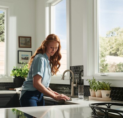 A woman wiping down counters in a sunny kitchen with light pouring through three large V450 windows
